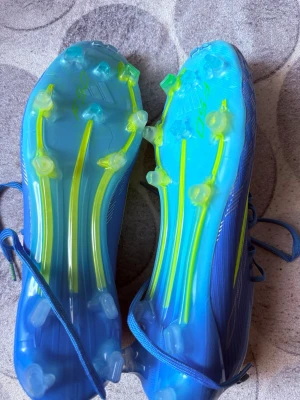 Blåa Adidas X fotbollsskor F50 - Säljer ett par blåa Adidas X fotbollsskor med neon-gula detaljer och klassiska tre ränder på sidan. Skorna har snörning och en lätt ovandel i syntetmaterial samt FG-dobbar för spel på naturgräs. Perfekta för dig som vill ha speed och kontroll på planen.