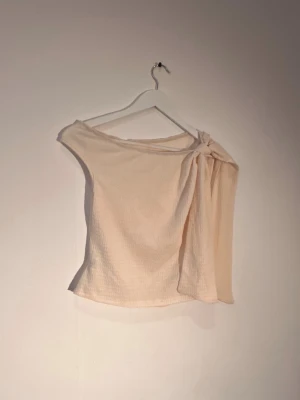 Beige oneshoulder topp med knyt - Trendig beige oneshoulder topp med snygg knyt-detalj på axeln. Tillverkad i ett mjukt, lätt strukturerat tyg som ger en avslappnad men stilren look. Perfekt för dig som vill sticka ut med en unik topp i garderoben.
