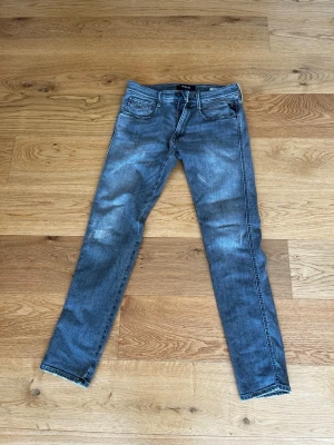 Grå slim fit jeans från Replay - Snygga grå slim fit jeans från Replay med klassisk femficksdesign och dragkedjegylf. Jeansen har en tvättad look med ljusare partier och svarta detaljer vid knappar och etikett. Perfekta för en trendig och avslappnad stil.  W33 L32