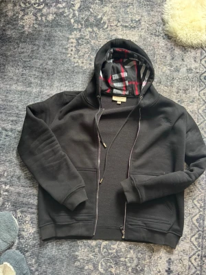 Svart hoodie från Burberry med dragkedja - Svart hoodie från Burberry med dragkedja och klassiskt rutigt foder i huvan. Hoodien har dragsko i huvan, ribbade muddar och två fickor framtill. Perfekt för en stilren och avslappnad look.