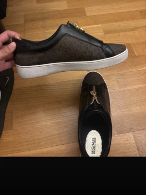 Bruna Michael Kors sneakers med dragkedja - Snygga bruna sneakers från Michael Kors med svart läderdetalj och guldfärgad dragkedja på ovansidan. Skorna har vit sula och klassiskt MK-mönster på sidan. Perfekta för dig som gillar stilrena och trendiga skor.