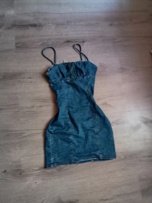 Kort jeansklänning med smala axelband - Snygg kort jeansklänning med smala axelband och markerad midja. Klänningen har dragkedja i ryggen och är figurnära i modellen. Perfekt för dig som gillar en trendig och avslappnad stil.