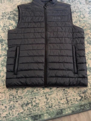 Svart dunväst från Old Navy - Stilren svart dunväst från Old Navy med horisontella sömmar och hög krage. Västen har två fickor med dragkedja och stängs med en hel dragkedja framtill. Perfekt för lager-på-lager och enkel att matcha med olika outfits.