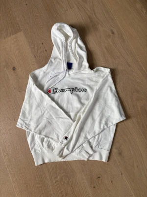 Vit hoodie från Champion - Säljer en fräsch vit hoodie från Champion med klassisk logga broderad på bröstet. Hoodien har huva med dragsko och ribbade muddar. Perfekt för en chill och sportig stil. Materialet är mjukt och har inga tecken på slitage 