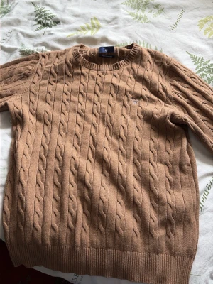 Beige kabelstickad tröja Gant - Kabelstickad tröja i beige från Gant med rund halsringning och diskret logga på bröstet. Tröjan har långa ärmar och ribbstickade muddar vid ärmslut och nederkant. Perfekt för lager-på-lager och snygg till jeans.