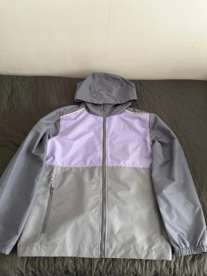 Under Armour Vindjacka  - Snygg vindjacka från Under Armour i grått och lila. Jackan har huva, hel dragkedja framtill och två sidofickor med dragkedja. Tillverkad i lätt och vindtätt material, perfekt för blåsiga dagar. Under Armour-logga på bröstet.  Storlek XS 