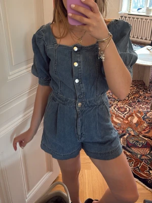 Blå jeansjumpsuit med puffärm - Snygg jeansjumpsuit i blå denim med korta ben och puffärmar. Knäppning framtill med silvriga knappar och markerad midja. Trendig fyrkantig ringning och avslappnad passform. Perfekt för dig som gillar en cool och avslappnad stil.