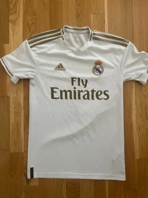 Real Madrid matchtröja 2020 - Snygg vit Real Madrid matchtröja från Adidas med gulddetaljer och klubbmärke på bröstet. Tröjan har Fly Emirates-tryck och är tillverkad i lätt och ventilerande Climalite-material. Perfekt för fotbollsträning eller att bära på läktaren.