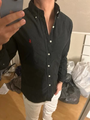 Svart skjorta från Ralph Lauren - Klassisk svart skjorta från Ralph Lauren med vit knappstängning och röd broderad logga på bröstet. Skjortan har lång ärm, button-down krage och är tillverkad i bomull. Perfekt för en stilren look.