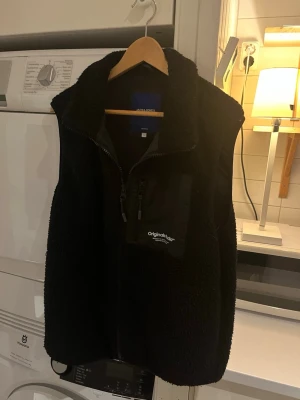 Svart fleeceväst från Jack & Jones - Mysig svart fleeceväst från Jack & Jones med dragkedja framtill och bröstficka med blixtlås. Västen har hög krage och är gjord i mjukt teddy-material. Perfekt att slänga över en hoodie eller tröja för extra värme.