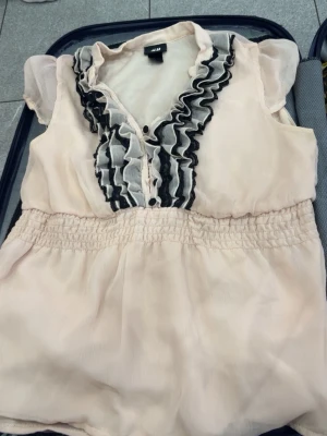 Ljusrosa blus med volang från H&M - Supersöt ljusrosa blus från H&M med korta puffärmar och volangdetaljer i svart och vitt längs knäppningen. Blusen har smock i midjan för en snygg siluett och är gjord i ett tunt, lätt material som känns luftigt och skönt.