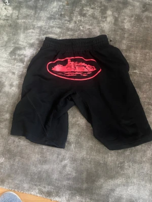 Svarta corteiz shorts - Svarta corteiz shorts med elastisk midja och ett stort rött tryck på baksidan. Shortsen har en avslappnad passform och är tillverkade i mjukt bomullsmaterial. Perfekta för en chill och bekväm stil.