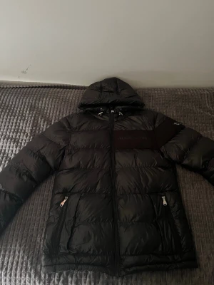 Svart pufferjacka från Alessandro - Snygg svart pufferjacka från Alessandro med huva och dragkedja framtill. Jackan har två framfickor med dragkedja och en tydlig logotyp på ärmen. Perfekt för kalla vinterdagar tack vare det vadderade materialet.