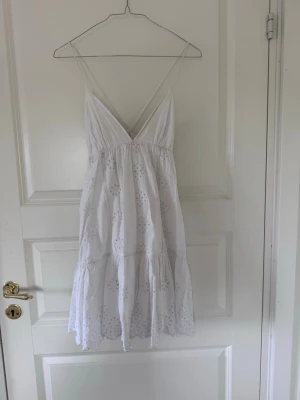 Vit somrig klänning med broderi - Superfin vit klänning med tunna axelband och v-ringning. Klänningen har broderade detaljer och volang nertill, samt snörning i ryggen. Perfekt för varma dagar och har en luftig passform.