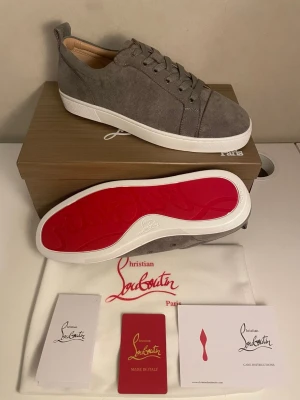 Gråa Christian Louboutin sneakers - Snygga gråa sneakers från Christian Louboutin i mocka med klassisk röd sula och vita kanter. Skorna har lågt skaft, rund tå och matchande grå snörning. Perfekta för dig som vill ha en exklusiv och stilren look med en ikonisk touch.