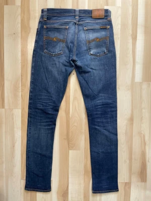 Blå slim fit jeans från Nudie Jeans - Snygga blå slim fit jeans från Nudie Jeans med klassisk femficksdesign och orangea sömmar. Jeansen har en medelhög midja och är tillverkade i denim med lätt tvättad look. Perfekta för en avslappnad och trendig stil.