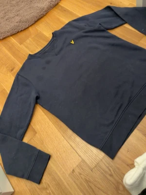 Mörkblå sweatshirt från Lyle & Scott - Mörkblå sweatshirt från Lyle & Scott med klassisk rund hals och gul logga broderad på bröstet. Tröjan har långa ärmar och ribbade muddar vid ärmslut och nederkant. Tröjan står att den är från 12-13 år men är i passform S-M. Nypris 900kr.