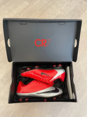 Nike CR7 Mercurial fotbollsskor röd/silver - Snygga Nike CR7 RGN fotbollsskor i rött och silver med svart swoosh. Perfekta för dig som vill sticka ut och prestera på topp. De är aldrig använda enbart testade på en mjuk matta.