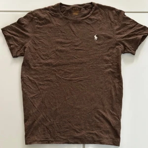 2 för 1 på Brun t-shirt från Polo Ralph Lauren - Brun t-shirt från Polo Ralph Lauren med klassisk vit broderad logga på bröstet. Modellen är Custom Slim Fit och har rund hals och korta ärmar. Tillverkad i mjuk bomull som känns skön mot huden. Perfekt för en avslappnad och stilren look.