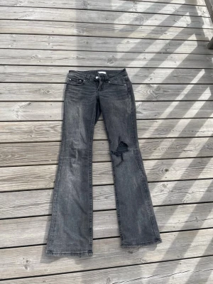 Grå bootcut jeans med slitning - Säljer ett par grå bootcut jeans med slitning på ena benet och stora bakfickor med knappdetaljer. Jeansen har låg midja och klassisk femficksdesign. Perfekta för en avslappnad och trendig look.
