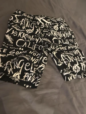 Feta svarta shorts inför sommaren - Coola svarta shorts med vit graffiti-inspirerad text och tryck över hela plagget. De har elastisk midja och avslappnad passform, perfekta för en avslappnad streetwear-look. Materialet känns lätt och bekvämt, perfekt för varma dagar.
