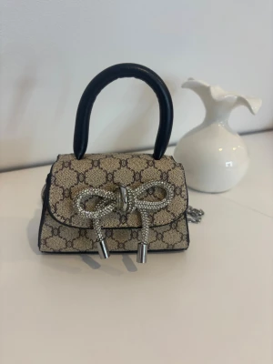 Beige miniväska med kedja - Superfin beige miniväska med klassiskt mönster och svart kant. Väskan har ett dekorativt silvrigt rosettspänne framtill och en lång silverfärgad kedja för crossbody. Perfekt accessoar för att lyfta din outfit.