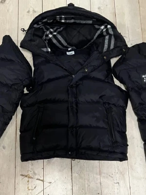Svart dunjacka från Burberry - Snygg svart dunjacka från Burberry med avtagbara ärmar så den kan användas som väst. Jackan har dragkedja, tryckknappar och två fickor framtill. Insidan av huvan har klassiskt Burberry-rutmönster i grått och vitt.