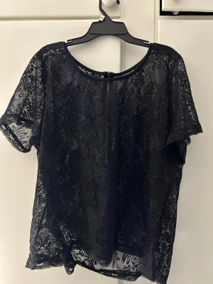 Svart spetsblus med dragkedja - Snygg svart blus i spets med korta ärmar och rund halsringning. Blusen har en dekorativ dragkedja i ryggen och är genombruten i materialet, vilket ger en cool och trendig look.