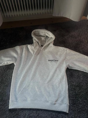 Grå Essentials Fear of God hoodie - Snygg grå hoodie från Essentials Fear of God med svart logga på bröstet. Klassisk känguruficka framtill och huva med dragsko. Perfekt för en chill och stilren look.