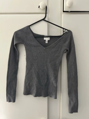 Grå ribbad v-ringad topp H&M - Snygg grå ribbad topp från H&M med v-ringning och lång ärm. Modellen är figurnära och har en stilren look som passar till många outfits. Perfekt för dig som gillar enkla men trendiga plagg.