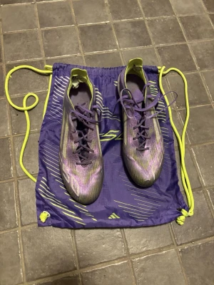 Lila Adidas X crazyfast fotbollsskor elite - Säljer ett par lila Adidas X crazyfast elite fotbollsskor med gula detaljer och snörning. Skorna har en smal, aerodynamisk form och är tillverkade i syntetmaterial för lätt vikt och bra bollkontroll. Medföljer matchande gympåse i lila och gult.