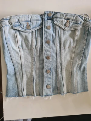 Ljusblå jeanskorsett med knappar - Trendig ljusblå korsettopp i denim med knappar framtill och två dekorativa fickor upptill. Snygga sömdetaljer och rå kant nertill ger en cool vibe. Perfekt att styla med höga jeans eller kjol.