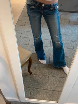 Blå bootcut jeans med snygg design på fickorna - Säljer ett par blå bootcut jeans med slitningar på knäna och snygga detaljer på bakfickorna. Jeansen har låg midja och klassisk femficksdesign. Perfekta för dig som gillar en avslappnad och trendig stil.