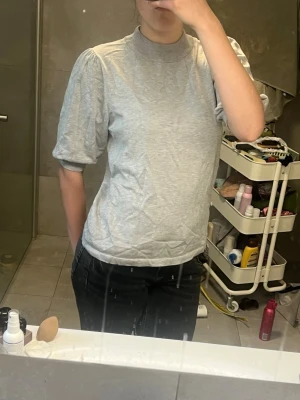 Grå topp med puffärm och hög krage - Snygg grå topp med puffiga kortärmar och hög ribbad krage. T-shirten har en loose passform och är tillverkad i mjukt material som känns skönt mot huden. Perfekt att styla med jeans eller kjol för en trendig look.