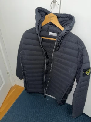 Svart pufferjacka från Stone Island - Snygg svart pufferjacka från Stone Island med ikonisk logotyp på ärmen. Jackan har huva, dragkedja framtill och quiltad design. Perfekt för kalla dagar och ger en stilren look. Materialet är lätt och isolerande för extra värme. Ganska slim fit jacka. Den är äkta köpt i NK Stockholm.