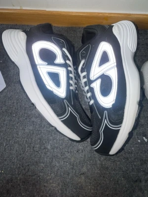 Svarta Dior sneakers med reflexdetaljer - Säljer ett par svarta Dior sneakers med vita och reflekterande detaljer. Skorna har vit sula, svart mesh och vita skosnören. Snyggt mönster på sidorna och modern chunky design. Perfekta för dig som gillar streetwear och vill sticka ut.