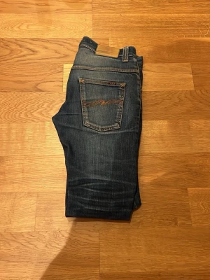 Nudie jeans grisch - Snygga mörkblå jeans från Nudie Jeans med klassisk femficksdesign och orange sömmar på bakfickan. Storlek 30/32.  Jeansen har en smal passform och är tillverkade i denim med lätt slitning för en cool look.