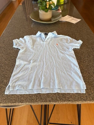 Ljusblå pikétröja från Ralph Lauren - Klassisk ljusblå pikétröja från Ralph Lauren med korta ärmar och krage. Tröjan har en liten broderad logga i orange på bröstet och är tillverkad i bomull. Perfekt för en avslappnad och stilren look.