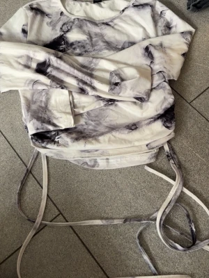 Vit och grå långärmad topp Shein - Trendig långärmad topp från Shein i vitt med grått mönster. Toppen har snörning i midjan som kan knytas för en snygg passform. Mjuk och lätt i materialet, perfekt till jeans eller kjol.