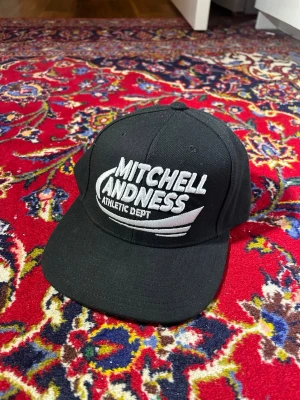 Svart keps från Mitchell & Ness - Snygg svart snapback-keps från Mitchell & Ness med broderad vit logga framtill och justerbar baksida. Klassisk rak skärm i grått och cool streetwear-vibe. Perfekt för dig som vill ha en stilren och sportig look.