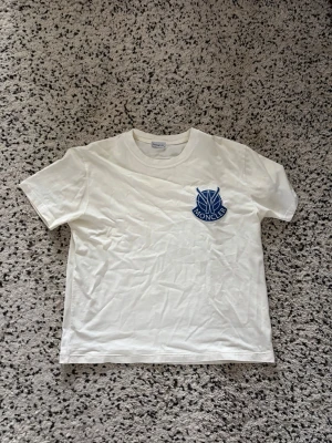 Vit Moncler t-shirt med blå logga - Snygg vit t-shirt från Moncler i storlek M. T-shirten har en blå Moncler-logga tryckt på vänster bröst och är gjord i mjuk bomull. Klassisk passform med rund hals och korta ärmar, perfekt till jeans eller shorts.