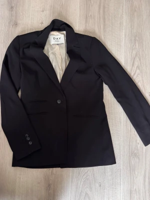 Svart kavaj från DAY Birger et Mikkelsen - Stilren svart kavaj från DAY Birger et Mikkelsen med klassisk passform och ett knappslå framtill. Kavajen har två fickor och ljus innerfoder. Perfekt för en snygg och tidlös look.