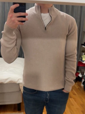 Beige stickad tröja från John Henric - Snygg beige stickad tröja med halvzip och ståkrage. Tröjan har ribbstickad struktur, långa ärmar och sitter tajt mot kroppen. Perfekt för lager-på-lager och passar till jeans eller chinos.
