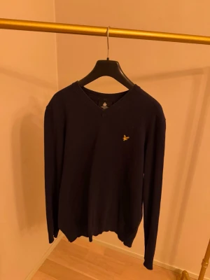 Mörkblå tröja från Lyle & Scott - Klassisk mörkblå tröja från Lyle & Scott med v-ringning och den ikoniska gula logon broderad på bröstet. Tröjan har lång ärm och är tillverkad i ett mjukt material som passar perfekt till många olika stilar.
