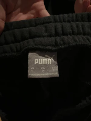 Svarta mjukisbyxor från Puma - Säljer ett par svarta mjukisbyxor från Puma med resår i midjan och smal passform. Byxorna har en vit Puma-logga på låret och är tillverkade i mjukt material.