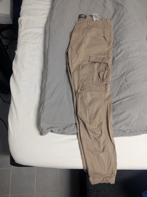 Beige cargobyxor från Jack & Jones - Säljer ett par beige cargobyxor från Jack & Jones med klassiska sidofickor och resår vid benslut. Byxorna har knapp- och dragkedjestängning samt flera praktiska fickor. Perfekta för en avslappnad och trendig stil. Använda 1 gång