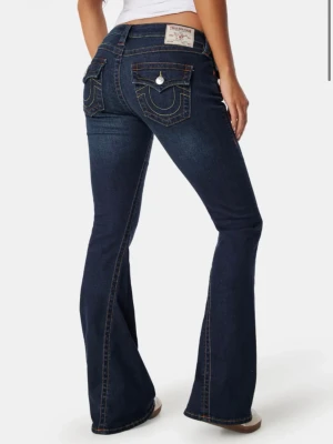 Mörkblå bootcut jeans True Religion - Säljer ett par mörkblå bootcut jeans från True Religion med klassiska kontrastsömmar och snygga fickdetaljer bak. Jeansen har låg midja och är tillverkade i stretchig denim för en bekväm passform. Perfekta för dig som gillar en tidlös och trendig look.