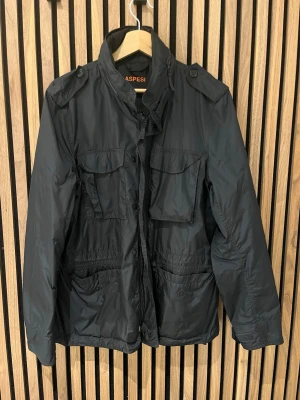 Blå vindjacka från Aspesi - Snygg svart vindjacka från Aspesi med fyra stora fickor framtill, hög krage och axelklaffar. Jackan har dragkedja och knappar samt en lätt glansig finish. Perfekt för dig som gillar stilren och funktionell design.