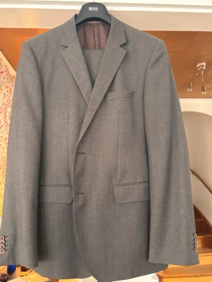 Snygg stilren kostym från Hugo Boss  - Stilren grå kostym från Hugo Boss i klassisk passform. Tyget har liv och karaktär – inte en tråkig, slätstruken grå, utan med subtila inslag av beige och bruna toner som ger ett mer levande och exklusivt intryck. Insidan har ett brunt foder med ett diskret rutmönster och eleganta beige detaljer.  Ett perfekt val för dig som vill ha en tidlös men samtidigt intressant och välklädd look – passar lika bra till arbete som till fest eller student. Mycket prisvärd. Nypris: 4 995 kr.
