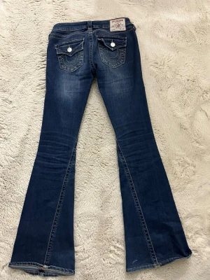 Low waist joey True Religion - Säljer ett par mörkblå bootcut jeans från True Religion strlk 25💕med snygga kontrastsömmar och ikoniska fickor med lock och vita knappar bak. Jeansen har en klassisk femficksmodell och är tillverkade i denim med lätt tvättad look.
